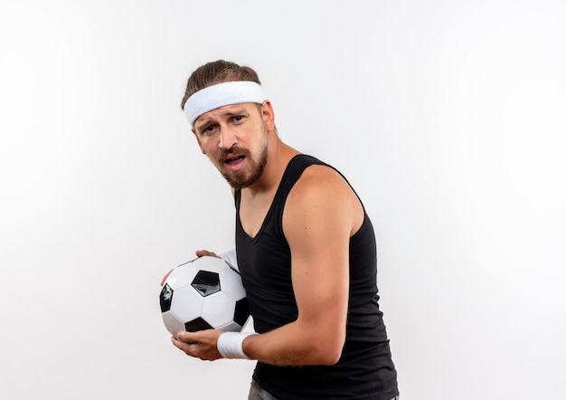 Jovem bonito e confiante homem desportivo usando bandana e pulseiras segurando uma bola de futebol, olhando com a boca aberta, isolada na parede branca com espaço de cópia