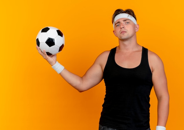 Jovem bonito e confiante homem desportivo usando bandana e pulseiras segurando uma bola de futebol, parecendo isolado em laranja