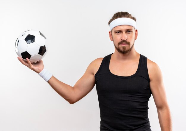 Jovem bonito e confiante homem desportivo usando bandana e pulseiras, segurando uma bola de futebol, parecendo isolado na parede branca
