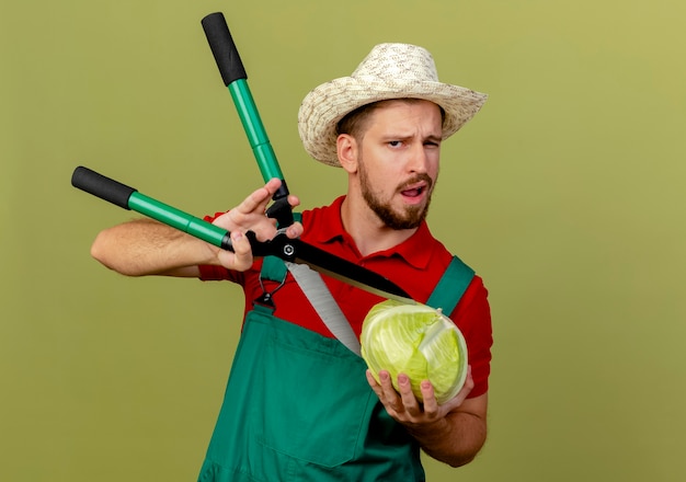 Foto grátis jovem bonito e confiante jardineiro eslavo de uniforme e chapéu segurando repolho e tentando cortá-lo com podadores isolados na parede verde oliva com espaço de cópia