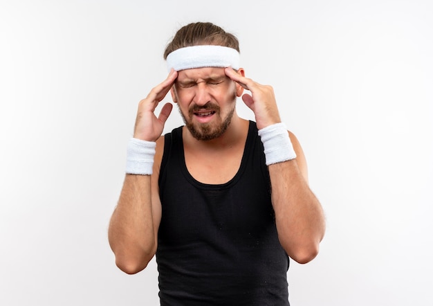 Jovem bonito e desportivo dolorido usando bandana e pulseiras, colocando as mãos nas têmporas, sofrendo de dor de cabeça com os olhos fechados, isolados na parede branca