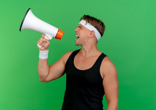 Jovem bonito e desportivo usando bandana e pulseiras, gritando no alto-falante, isolado em um fundo verde com espaço de cópia