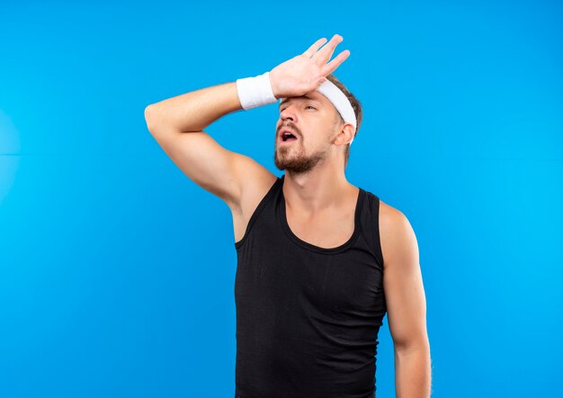 Jovem bonito e esportivo cansado usando bandana e pulseiras, colocando a mão na cabeça, olhando para o lado isolado no espaço azul