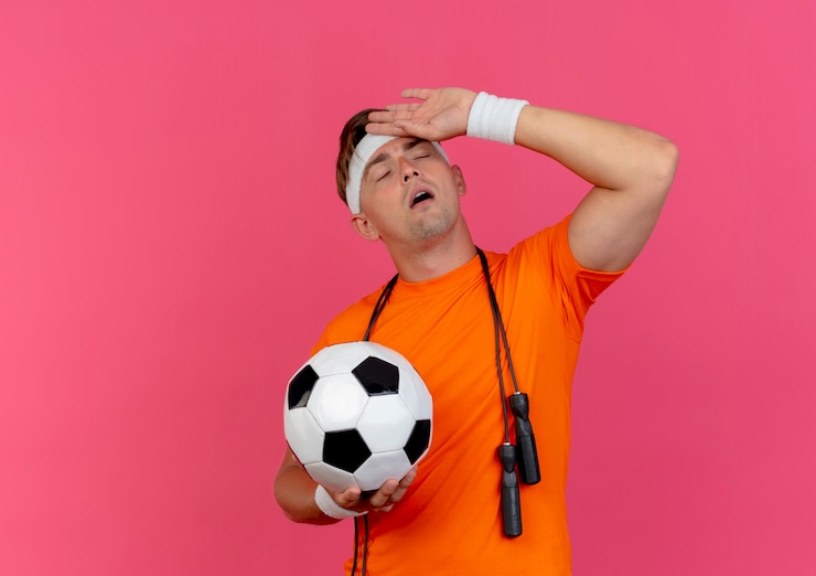 Foto grátis jovem bonito e esportivo cansado usando bandana e pulseiras com corda de pular em volta do pescoço, segurando uma bola de futebol, colocando a mão na testa com os olhos fechados, isolado no fundo rosa com espaço de cópia