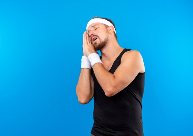 Jovem bonito e esportivo com sono usando bandana e pulseiras, fazendo gestos para dormir, isolado no espaço azul