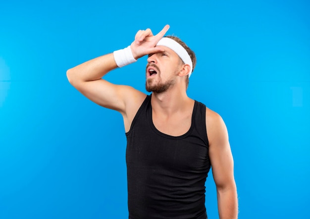 Jovem bonito e esportivo impressionado usando bandana e pulseiras, colocando a mão na testa e apontando para cima, isolado na parede azul