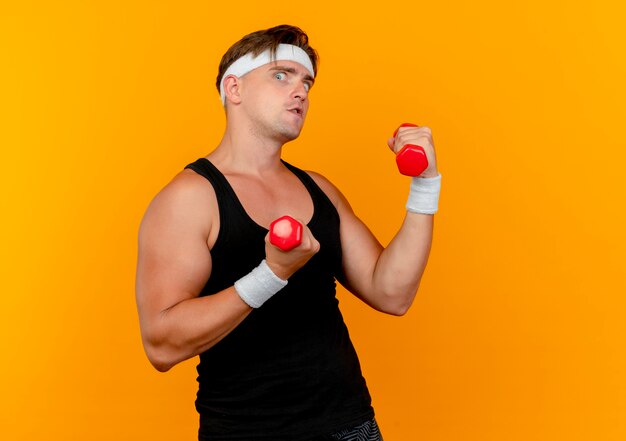 Jovem bonito e esportivo impressionado usando bandana e pulseiras segurando halteres isolados em laranja