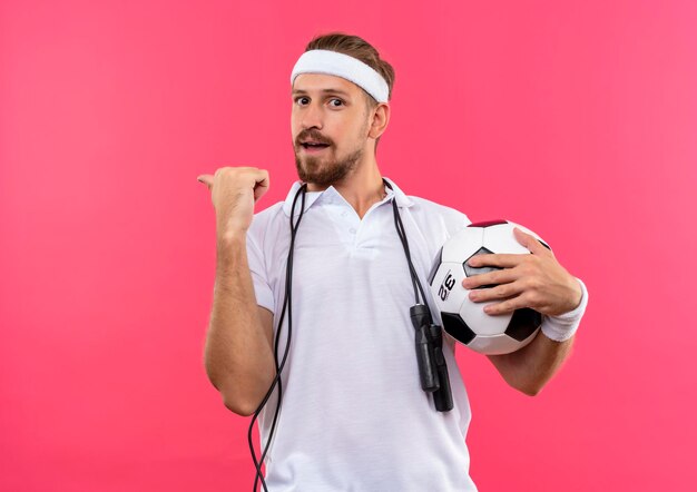 Jovem bonito e esportivo impressionado usando bandana e pulseiras segurando uma bola de futebol apontando para trás e com uma corda de pular em volta do pescoço isolada na parede rosa