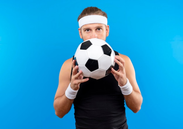 Jovem bonito e esportivo impressionado usando bandana e pulseiras, segurando uma bola de futebol e se escondendo atrás dela, isolado na parede azul