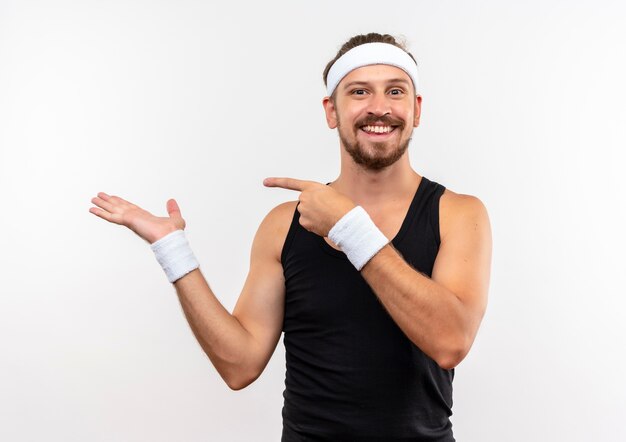 Jovem bonito e esportivo sorridente, usando bandana e pulseiras, mostrando a mão vazia e apontando para ela, isolado no espaço em branco