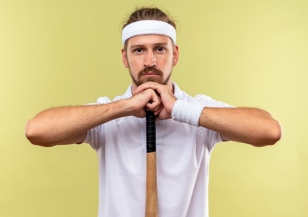 Jovem bonito e esportivo usando bandana e pulseiras segurando um taco de beisebol e colocando a cabeça nas mãos, parecendo isolado em um espaço verde