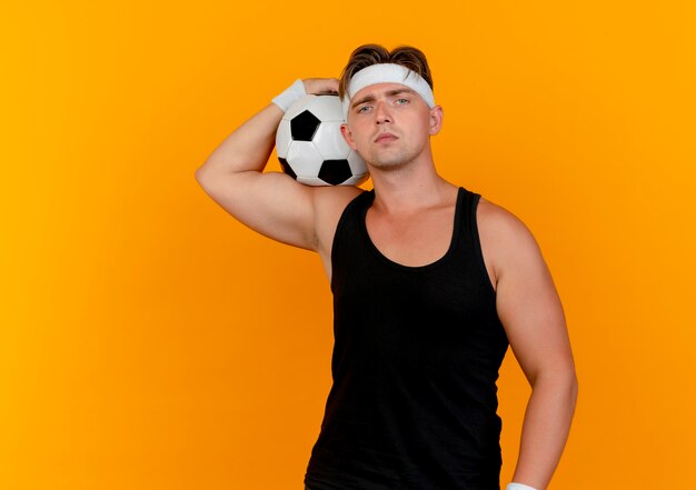 Jovem bonito e esportivo usando bandana e pulseiras segurando uma bola de futebol no ombro e olhando para a câmera isolada em um fundo laranja com espaço de cópia