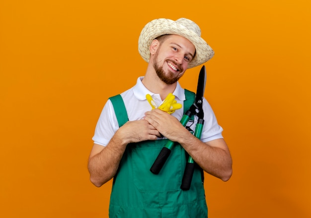 Jovem bonito e feliz jardineiro eslavo de uniforme, usando um chapéu, segurando luvas de jardinagem e podadores parecendo isolados