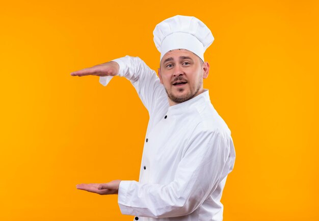 Jovem bonito e impressionado com uniforme de chef mostrando o tamanho isolado na parede laranja