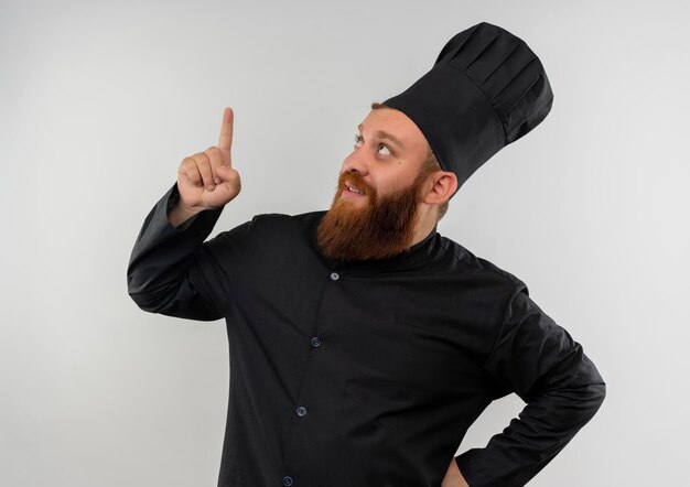 Jovem bonito e impressionado com uniforme de chef, olhando e apontando para cima, isolado na parede branca