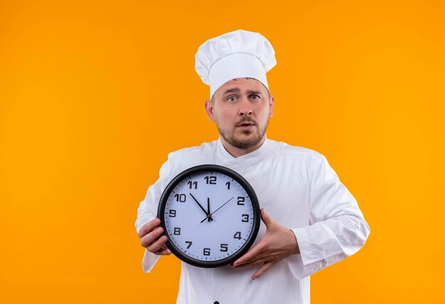 Jovem bonito e impressionado com uniforme de chef segurando o relógio isolado na parede laranja