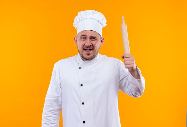 Jovem bonito e impressionado com uniforme de chef segurando o rolo de massa isolado na parede laranja