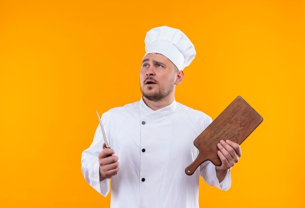 Jovem bonito e impressionado com uniforme de chef segurando uma faca e uma tábua de cortar, olhando para cima, isolado na parede laranja