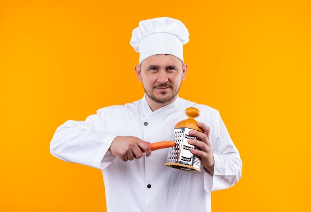 Jovem bonito e satisfeito com uniforme de chef ralando cenoura com ralador isolado em um espaço laranja