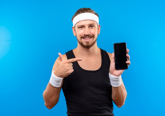 Jovem bonito e sorridente homem desportivo usando bandana e pulseiras segurando e apontando para um telefone celular isolado no espaço azul