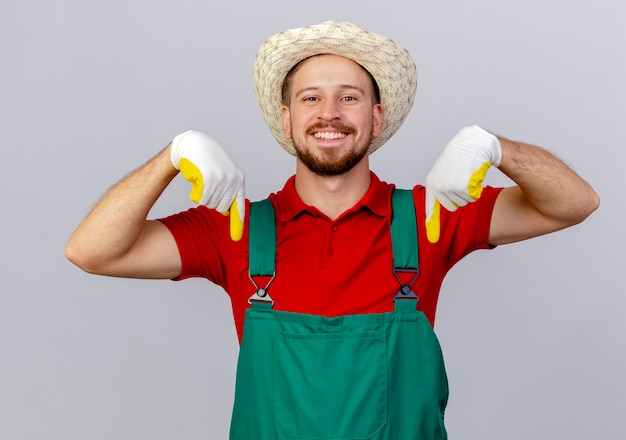 Jovem bonito e sorridente jardineiro eslavo de uniforme, usando luvas de jardinagem e chapéu apontando para baixo, isolado na parede branca