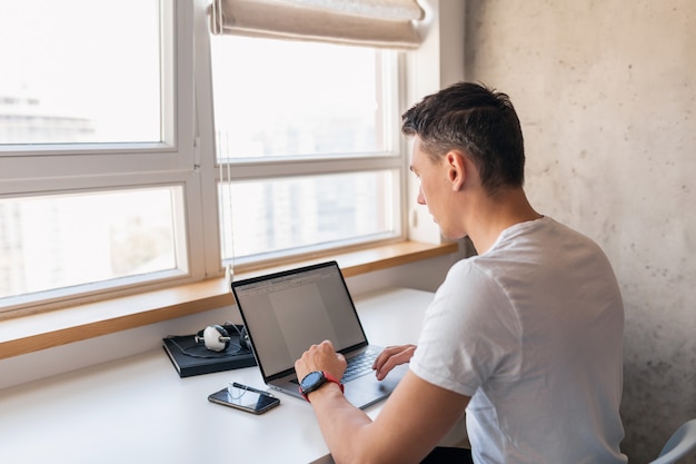 Jovem bonito em roupa casual sentado à mesa trabalhando no laptop, freelancer em casa