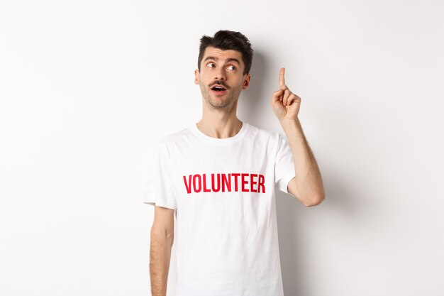 Jovem bonito em t-shirt de voluntário, tendo uma ideia, levantando o dedo e dizendo sugestão, fundo branco.