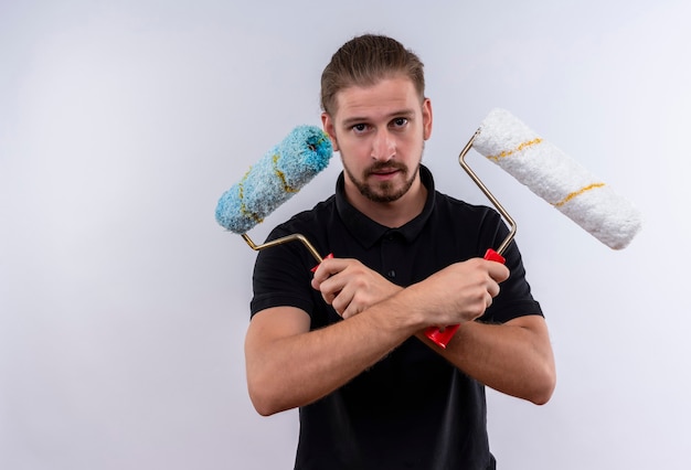 Foto grátis jovem bonito em uma camisa pólo preta em pé com os braços cruzados segurando rolos de tinta olhando para a câmera com uma cara séria em pé sobre um fundo branco