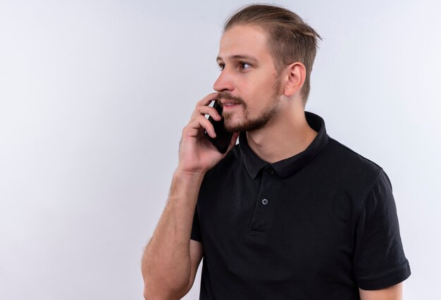 Jovem bonito em uma camisa pólo preta olhando para o lado sorrindo enquanto fala no celular em pé sobre um fundo branco