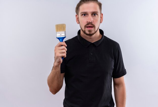 Jovem bonito em uma camisa pólo preta segurando um pincel e parecendo surpreso em pé sobre um fundo branco