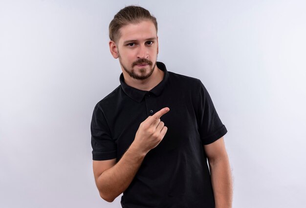 Jovem bonito em uma camisa pólo preta sorrindo confiante apontando para o lado com o dedo em pé sobre um fundo branco