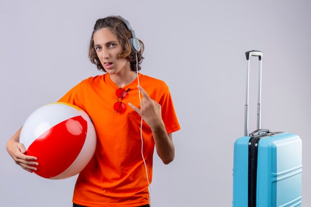 Jovem bonito em uma camiseta laranja com fones de ouvido segurando uma bola inflável, tornando o símbolo do rock, parecendo confiante em pé perto da mala de viagem sobre fundo branco