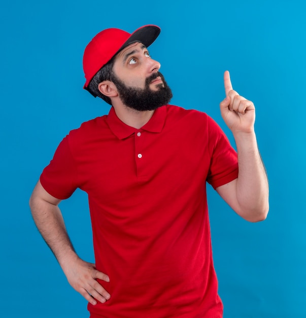 Foto grátis jovem bonito entregador caucasiano impressionado, vestindo uniforme vermelho e boné, colocando a mão na cintura, olhando e apontando para cima isolado no azul