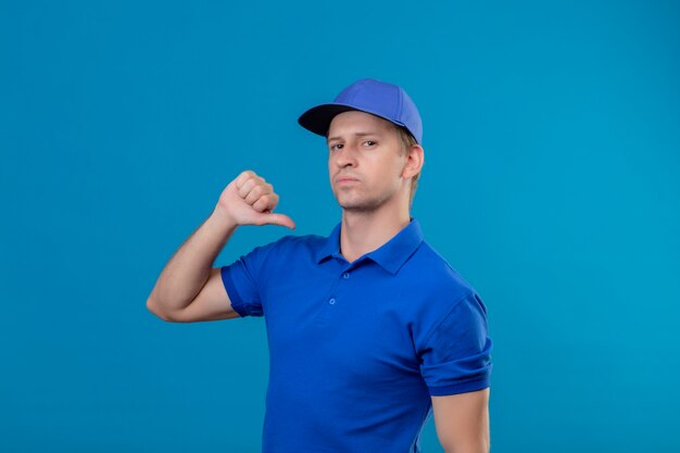 Jovem bonito entregador de uniforme azul e boné apontando para si mesmo com o polegar, parecendo confiante, satisfeito e orgulhoso de pé sobre a parede azul