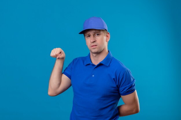 Jovem bonito entregador de uniforme azul e boné de punho cerrado, mostrando bíceps parecendo confiante em pé sobre a parede azul