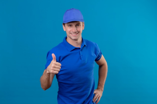 Jovem bonito entregador de uniforme azul e boné parecendo confiante sorrindo amigável mostrando os polegares em pé sobre a parede azul