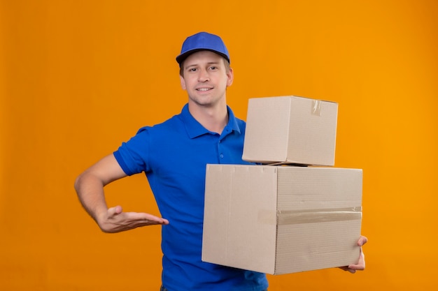 Jovem bonito entregador de uniforme azul e boné segurando caixas de papelão apresentando-se com o braço da mão em pé sobre a parede laranja