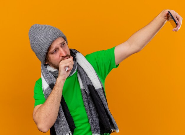 Jovem bonito eslavo doente usando chapéu de inverno e cachecol, tossindo, mantendo a mão na boca, tirando uma selfie isolada em um fundo laranja