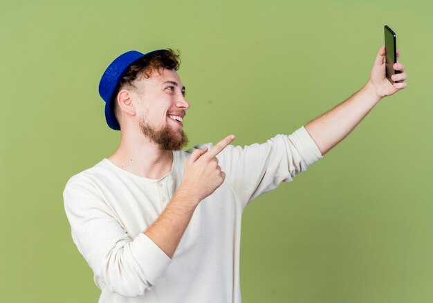 Jovem bonito eslavo festeiro alegre com chapéu de festa tomando selfie apontando para o celular isolado em fundo verde oliva