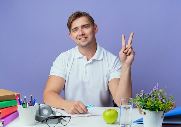 Jovem bonito estudante do sexo masculino satisfeito sentado na mesa com as ferramentas da escola, mostrando um gesto de paz isolado no azul