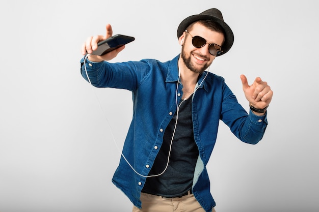 Jovem bonito feliz e sorridente ouvindo música em fones de ouvido isolados no fundo branco do estúdio, segurando o smartphone, vestindo camisa jeans, chapéu e óculos escuros