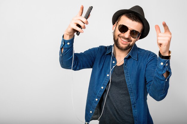 Jovem bonito feliz e sorridente ouvindo música em fones de ouvido isolados no fundo branco do estúdio, segurando o smartphone, vestindo camisa jeans, chapéu e óculos escuros