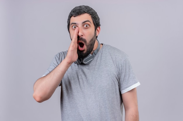 Jovem, bonito, homem, caucasiano, impressionado, usando fones de ouvido no pescoço, sussurrando isolado no branco com espaço de cópia