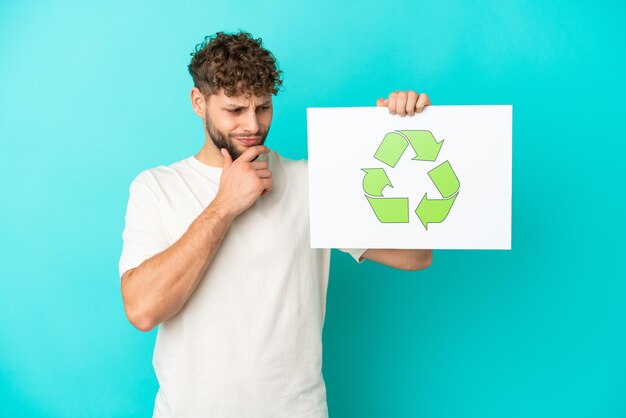 Jovem bonito homem caucasiano isolado em um fundo azul segurando um cartaz com o ícone de reciclagem