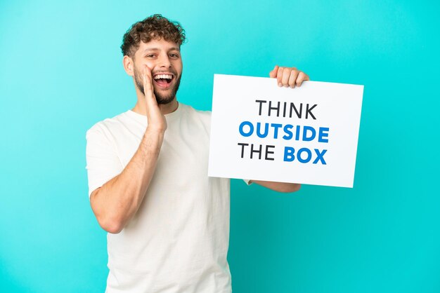 Jovem bonito homem caucasiano isolado em um fundo azul segurando um cartaz com o texto pense fora da caixa e gritando