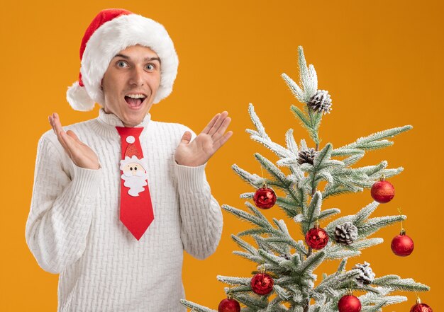 Jovem bonito impressionado com chapéu de Natal e gravata de Papai Noel em pé perto da árvore de Natal decorada, mostrando as mãos vazias isoladas na parede laranja