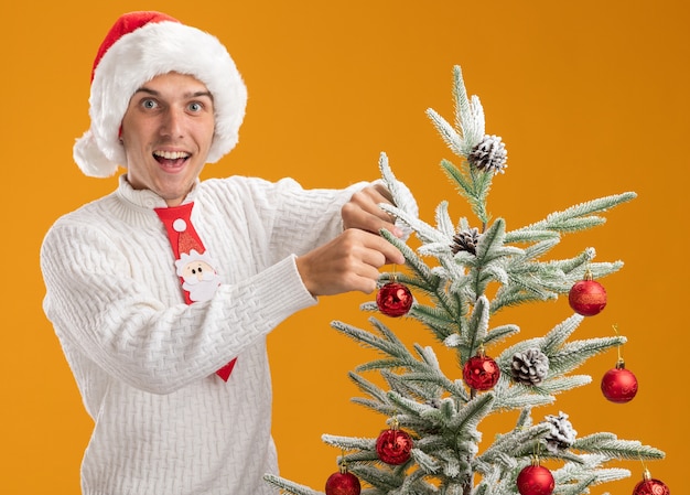 Jovem bonito impressionado com chapéu de natal e gravata de papai noel em pé perto da árvore de natal, decorando-o com enfeite de bola de natal isolado na parede laranja