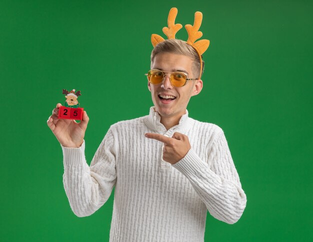 Jovem bonito impressionado com uma faixa de chifres de rena e óculos segurando e apontando para a árvore de Natal, brinquedo com data, olhando para a câmera isolada no fundo verde