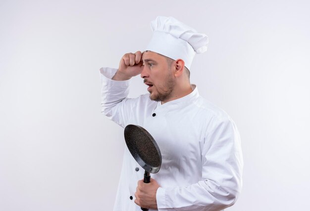 Jovem bonito impressionado com uniforme de chef segurando uma frigideira em pé na vista de perfil, olhando para o lado com a mão na cabeça isolada na parede branca