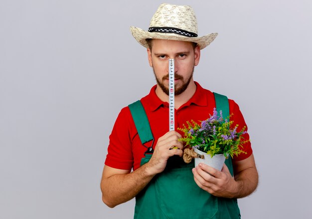 Jovem bonito jardineiro eslavo de uniforme e chapéu segurando fita métrica na frente do rosto olhando com um vaso de flores na outra mão isolado